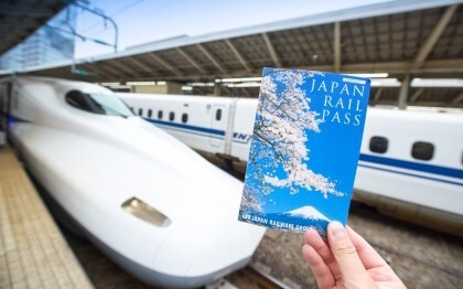 Eine Hand hält ein Japan Rail Pass Ticket vor einem Shinkansen im Bahnhof von Tokio